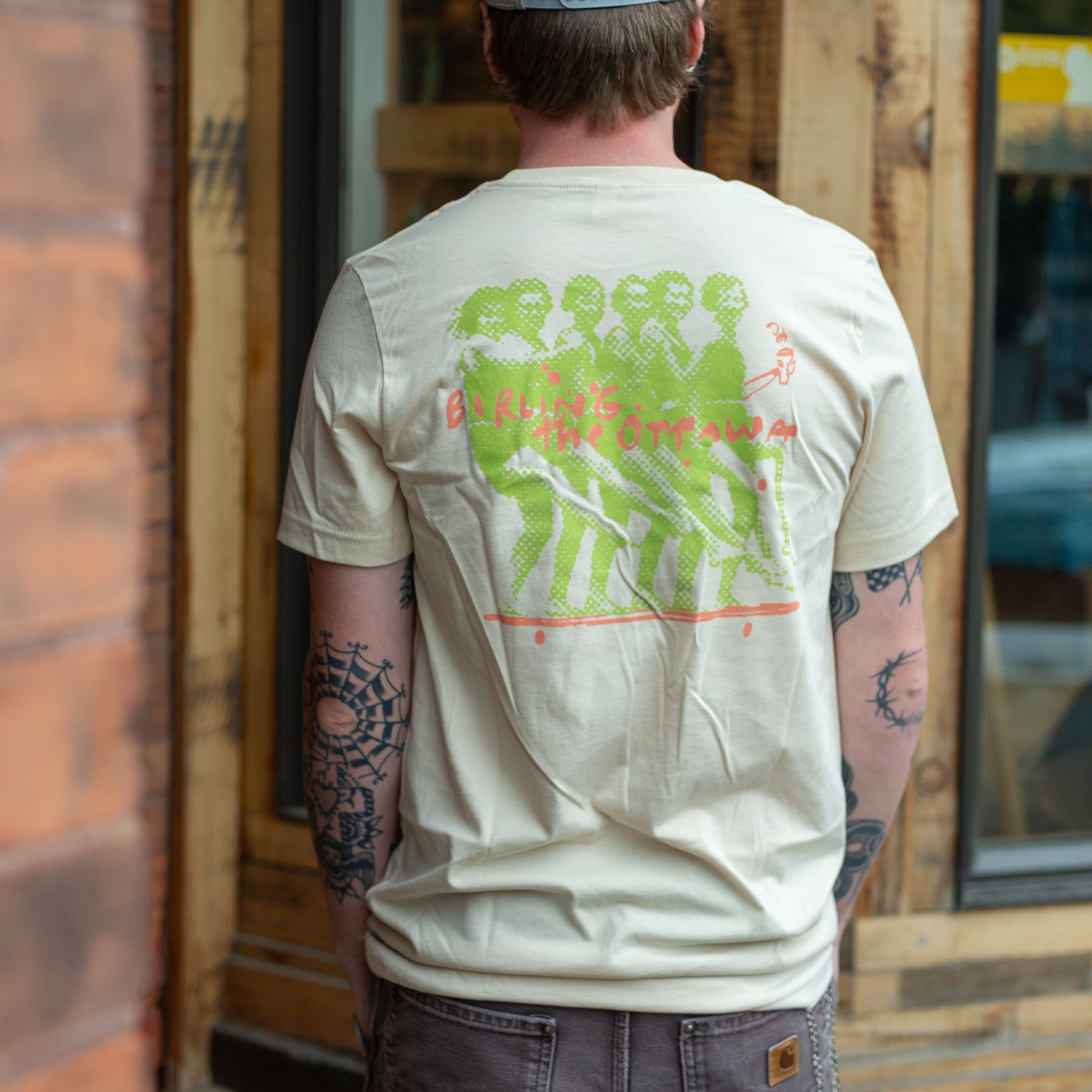 A man viewed from behind wearing a cream-colored t-shirt with a large green graphic print on the back, paired with dark gray cargo pants and a gray baseball cap, standing in front of a rustic wooden storefront.