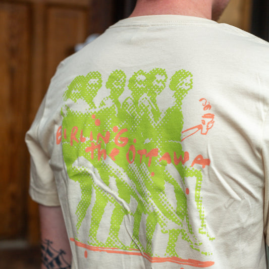 A man viewed from behind wearing a cream-colored t-shirt with a large green and orange graphic print on the back, set against a blurred wooden door background.