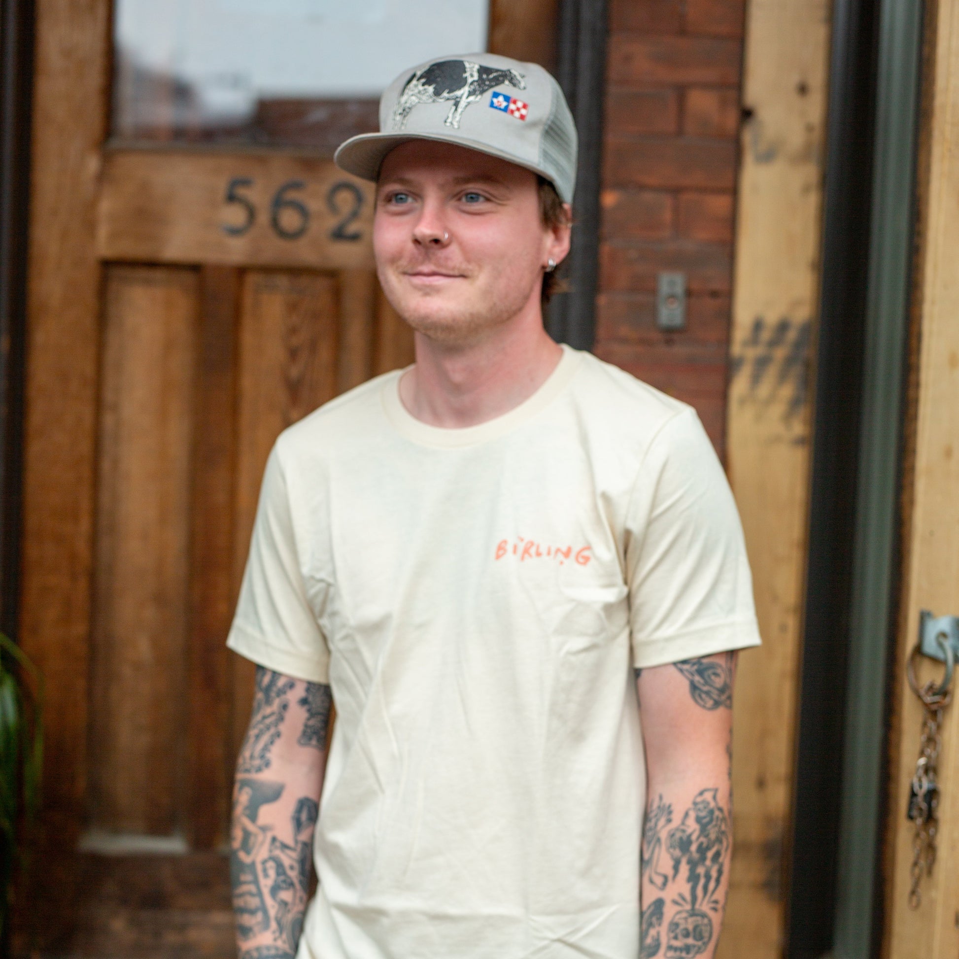 A man with tattoos stands in front of a wooden door wearing a cream-colored t-shirt with a small red graphic on the left chest, gray cargo pants, and a gray baseball cap.