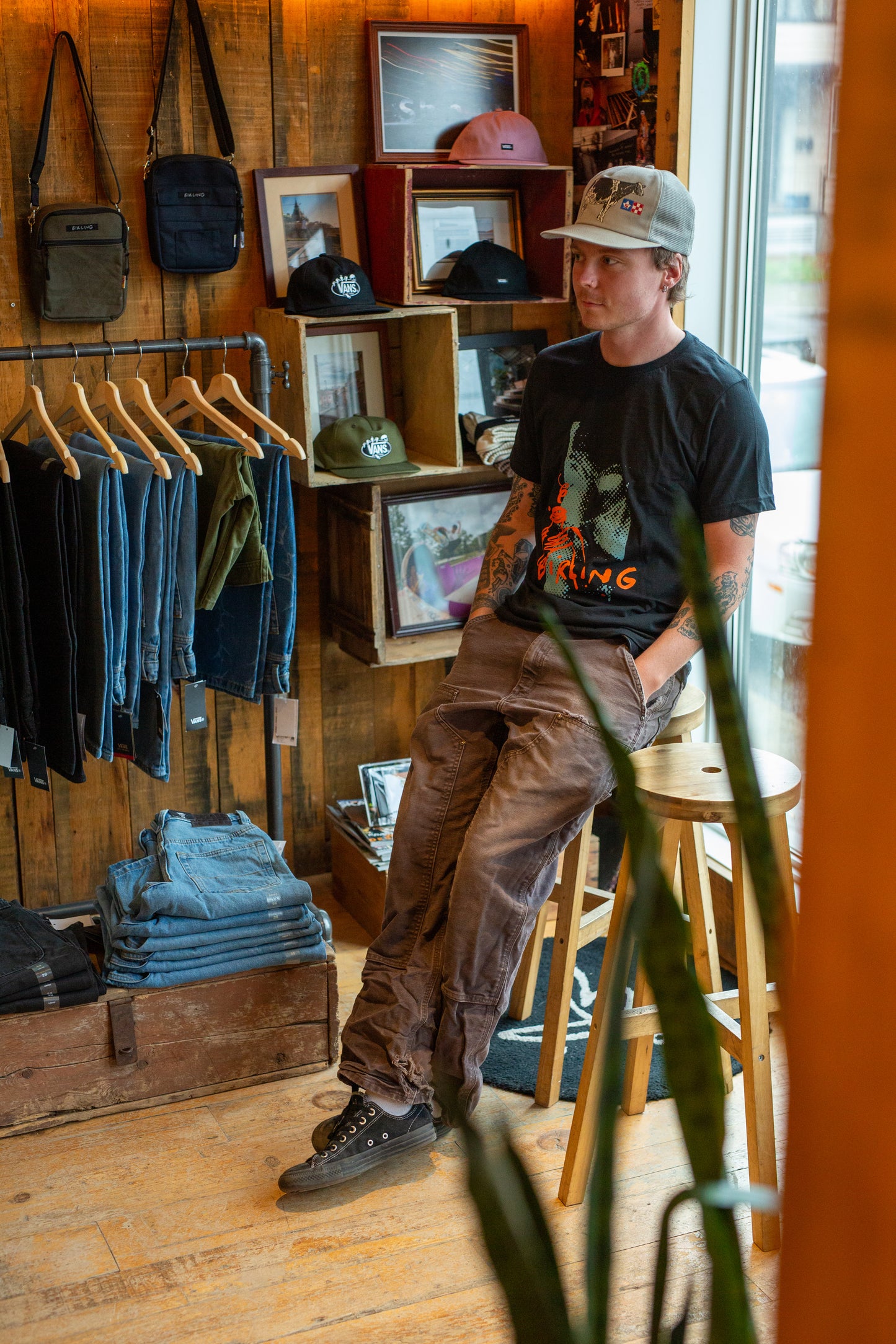 A man wearing a black graphic t-shirt, brown cargo pants, and a gray baseball cap sits on a wooden stool inside a rustic wood-paneled clothing store.
