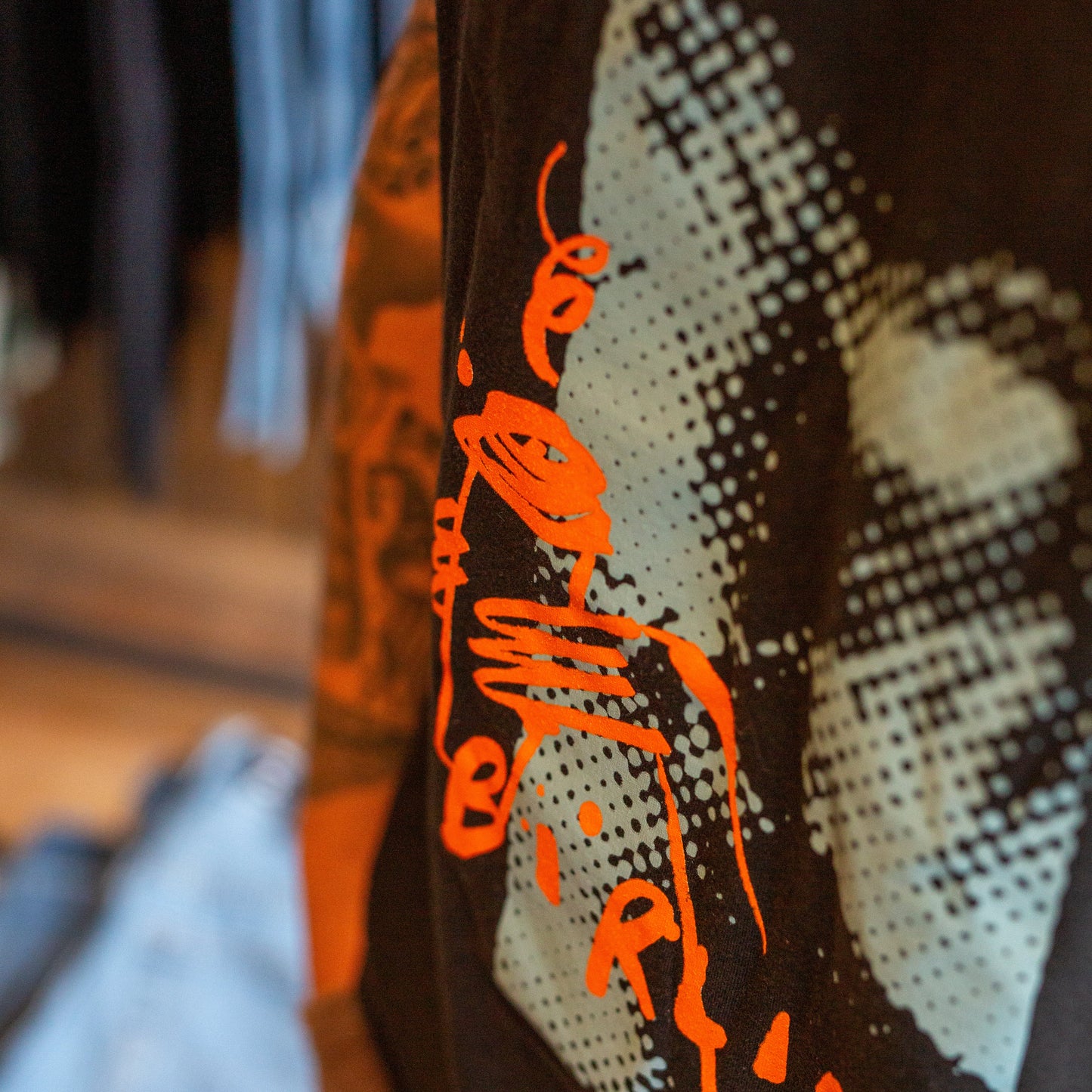 Close-up of a black graphic t-shirt featuring a large light gray halftone face print and bright orange stylized text, shown in a store setting.