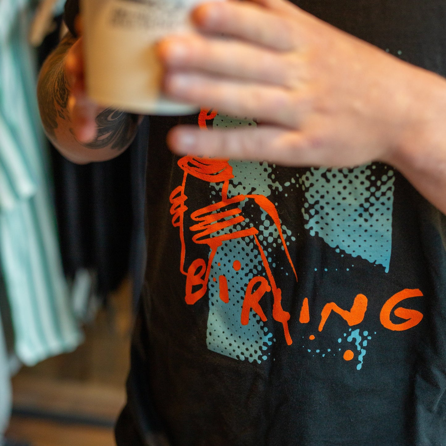 A person holding a white coffee cup in front of a black t-shirt with a graphic print. The graphic features orange text reading 'BIRING' and teal halftone shapes.