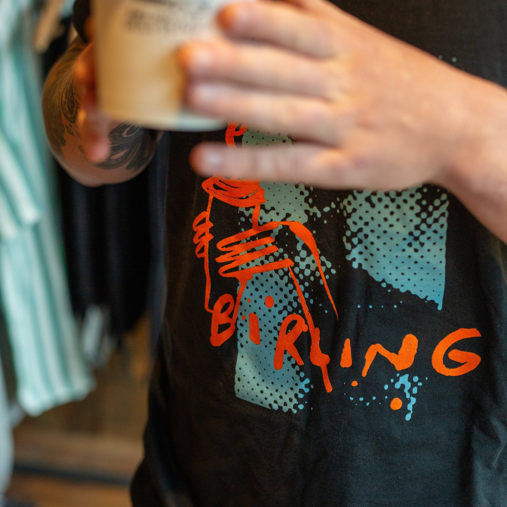 A person holding a white coffee cup in front of a black t-shirt with a graphic print. The graphic features orange text reading 'BIRING' and teal halftone shapes.
